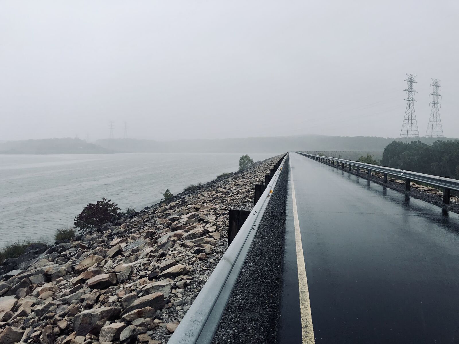 A wet road running along a rocky causeway into fog, with power towers in the distance
